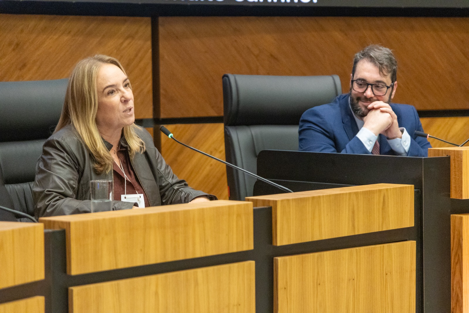 Maria Silvana é branca e loira. Usa camisa e casaco em tons de marrom e fala ao microfone na mesa oficial. A seu lado, Alexandre sorri com as mão cruzadas sobre a bancada da mesa oficial. Ele é branco, de cabelo e barba escuros. Usa camisa azul clara e blazer azul escuro, com óculos pretos de aro grande.