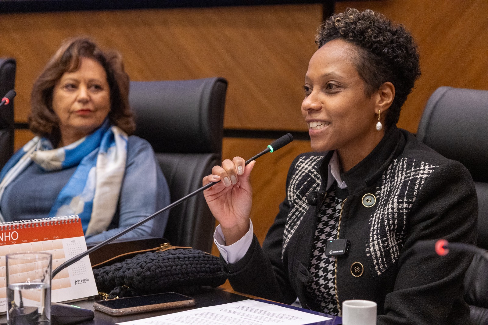 Leticia é negra, de cabelo preto curto. usa casaco xadrez e fala na mesa oficial. 