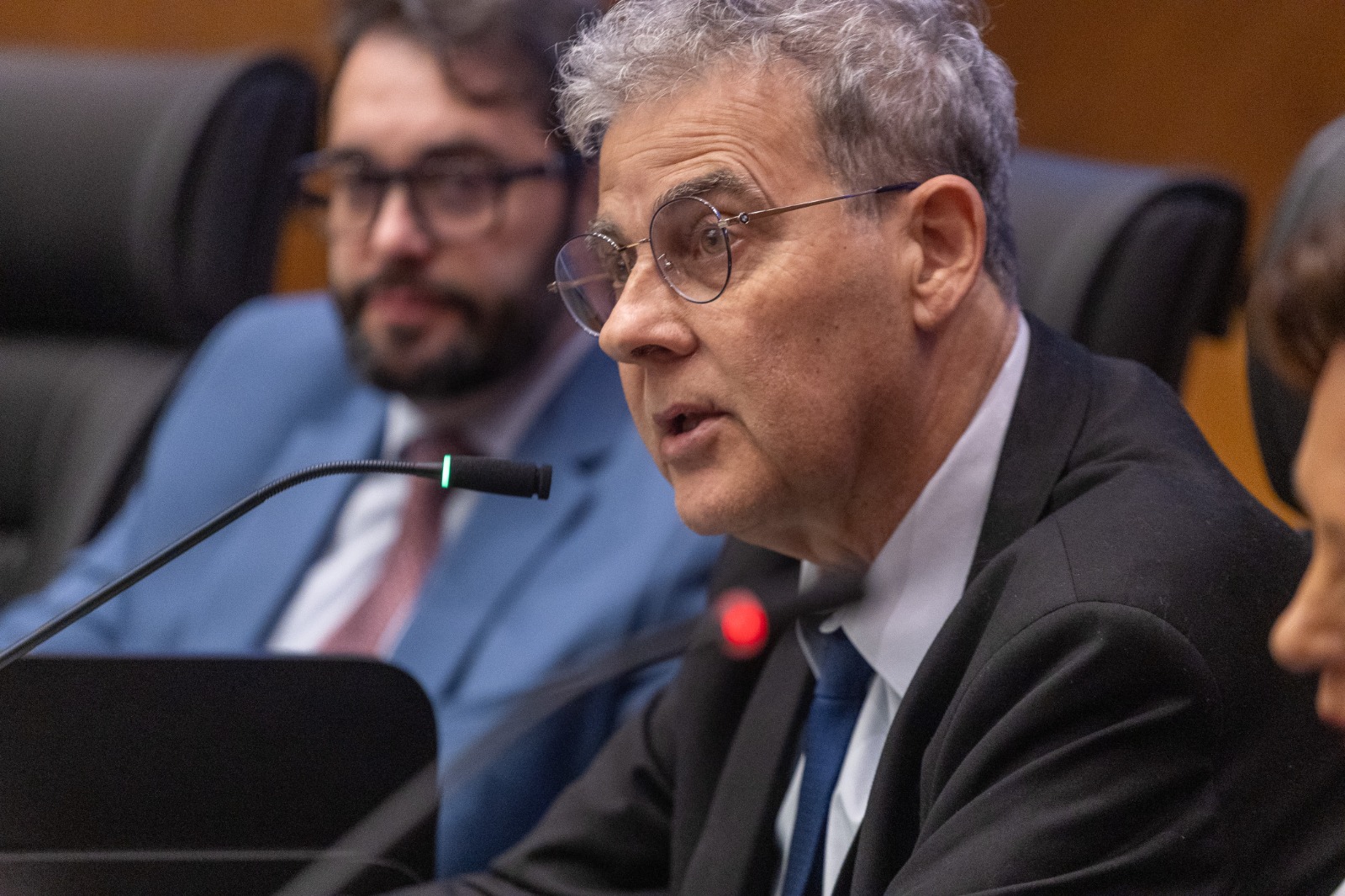 Ricardo é branco, de cabelo curto grisalho. usa óculos de aro redondo. usa terno cinza. 