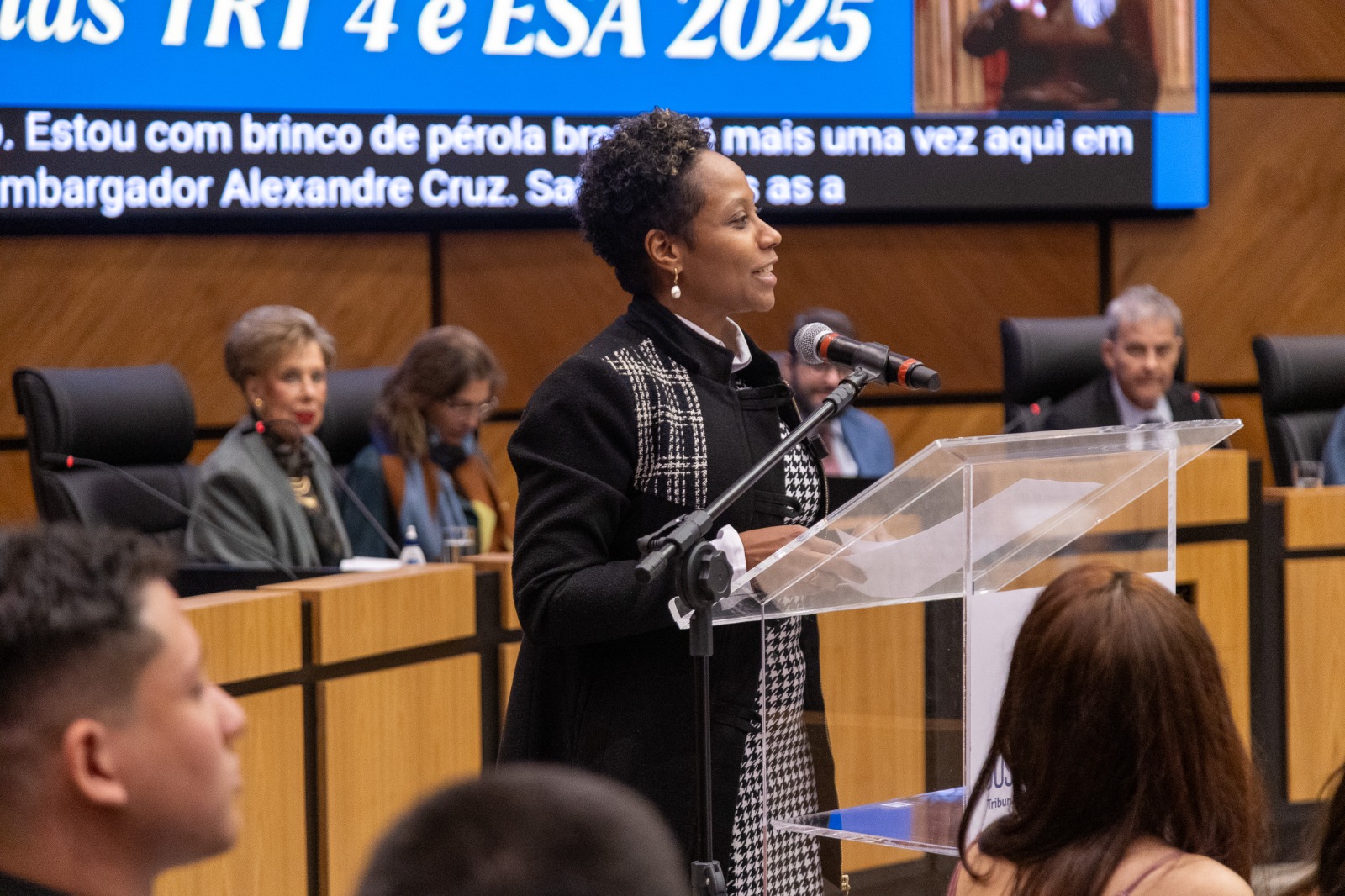 Leticia é negra, de cabelo preto e curto. Usa blazer preto com cinza. fala no púlpito. 