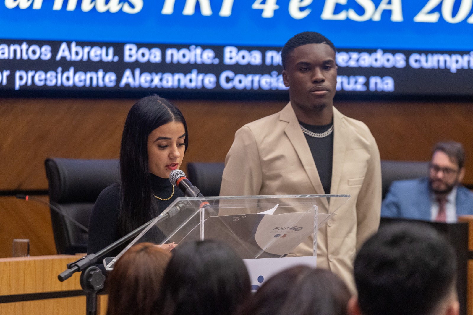 Ana Clara é branca, de cabelo liso escuro e usa blusa preta. Lucas é negro, de cabelo curto escuro e veste blazer creme.Os dois estão no púlpito.  