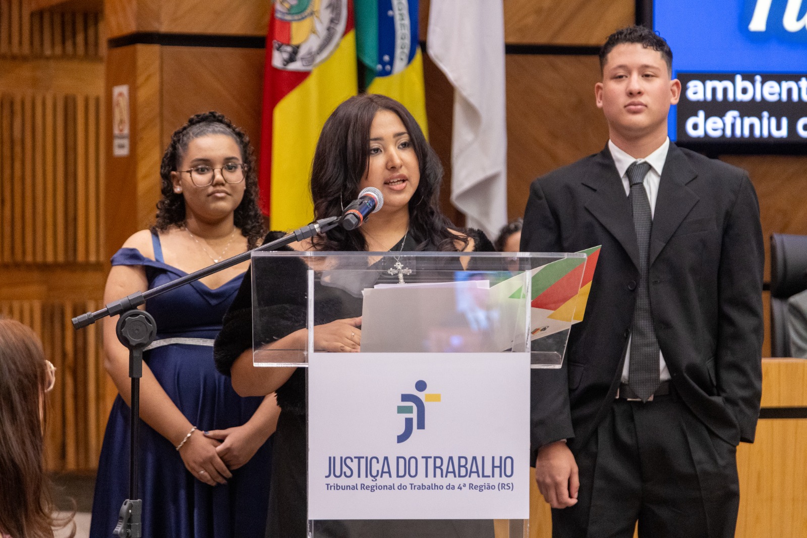 Thayssa é negra, usa cabelo cacheado preso e vestido azul. Anna está ao centro, é parda tem cabelo preto liso e usa vestido preto. José é branco, usa cabelo curto escuro, terno cinza e gravata cinza. 