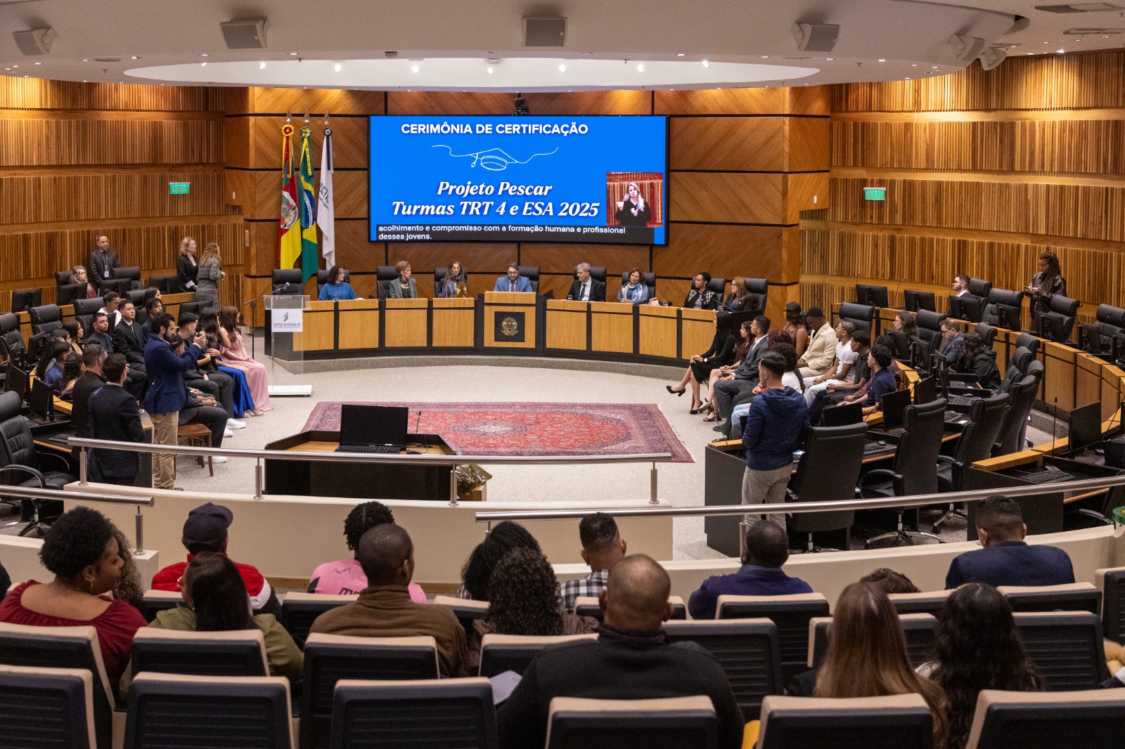 Foto geral do plenário com público em primeiro plano, de costas, e evento ocorrendo ao fundo, na nave. 