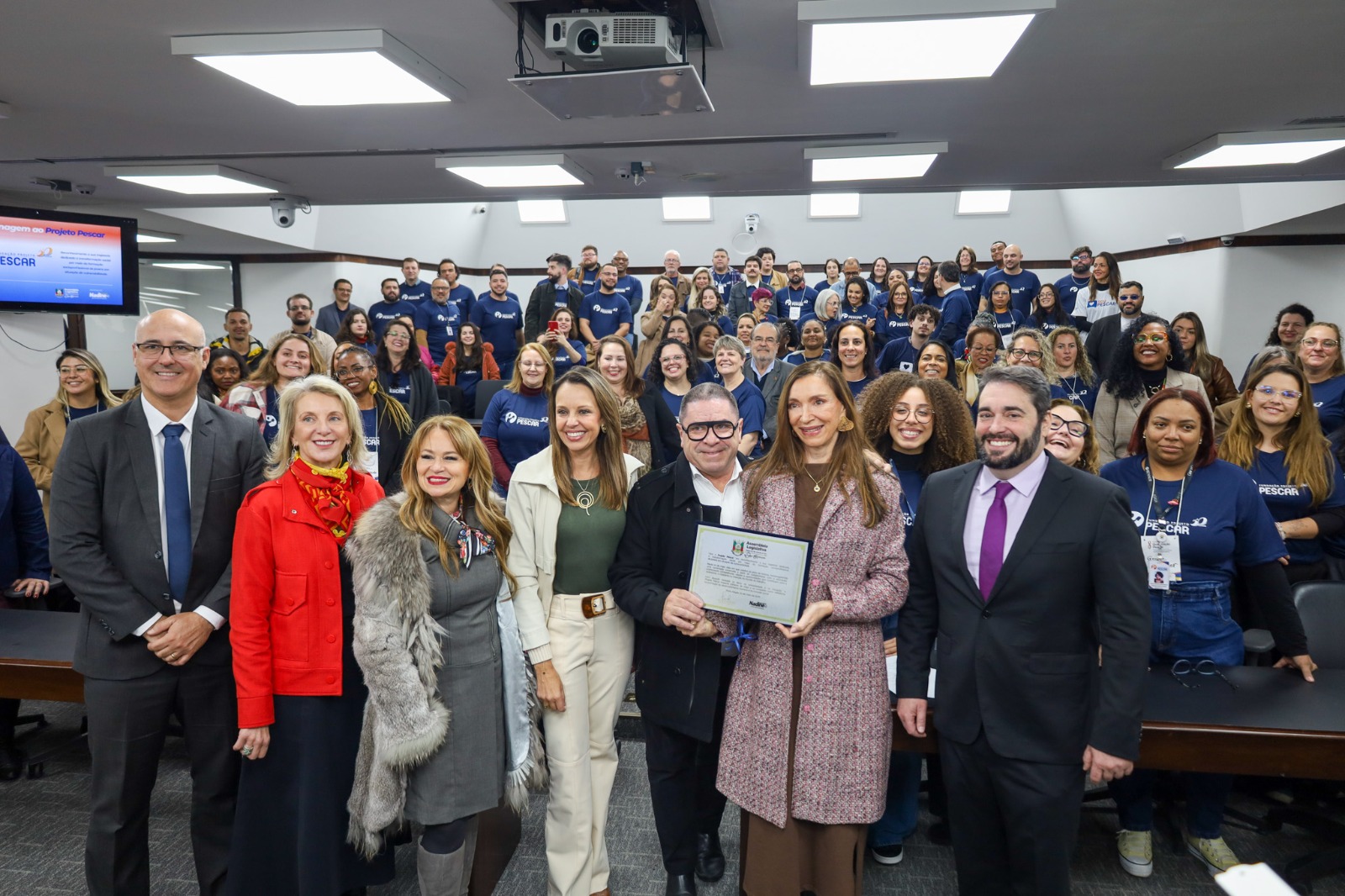 A foto mostra todos posando para foto com a placa em homenagem ao pescar.