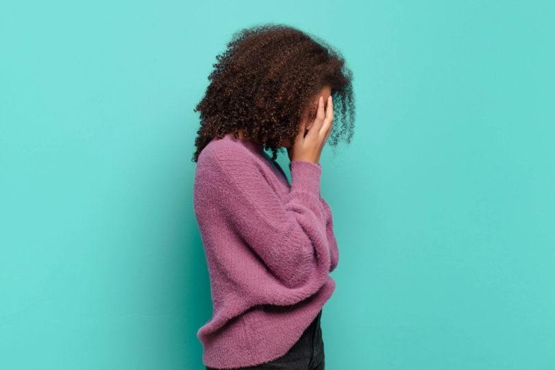 Jovem mulher negra tapa o rosto com as mãos, em demonstração de tristeza. Ela usa blusa lilás e o fundo é verde claro. Os cabelos são cacheados, pretos, pela altura dos ombros.