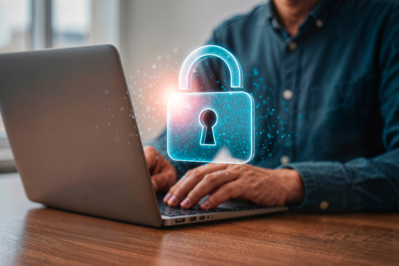 Homem digita em notebook. À frente, há a imagem de um cadeado, em azul. Conceito de confidenciaidade. Não há imagem do rosto e o homem usa camisa verde.