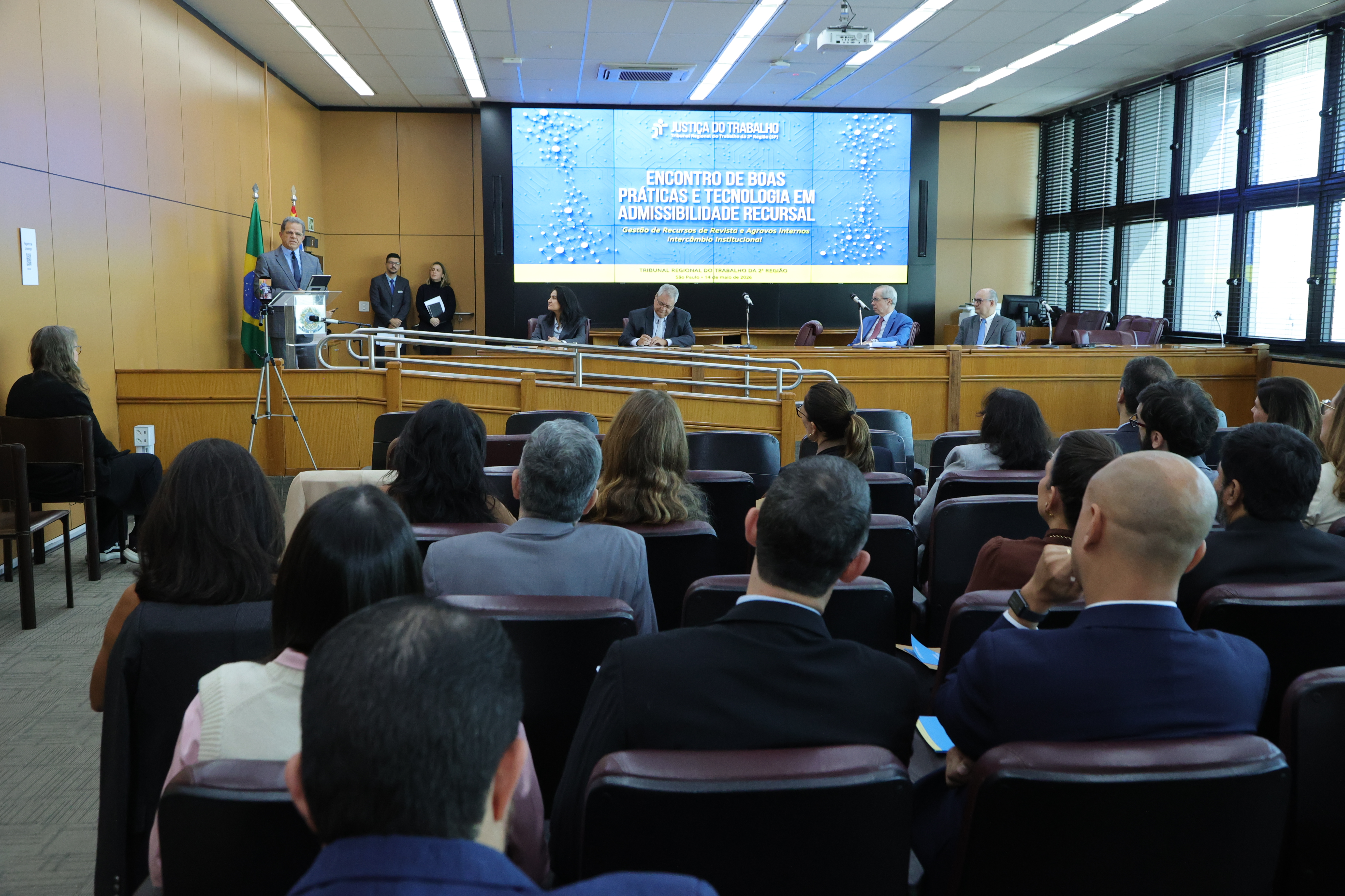 Foto em ângulo aberto, mostrando público em primeiro plano e mesa oficial ao fundo.