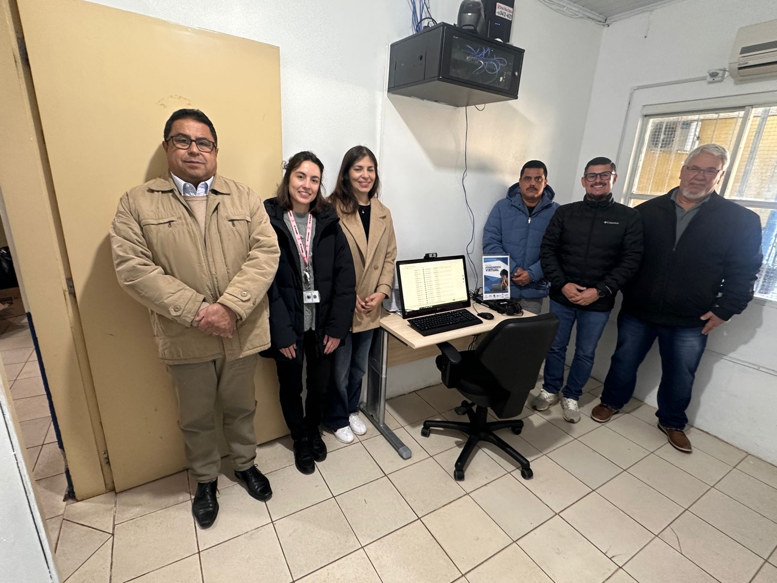 Seis pessoas posando em uma sala com piso claro, ao redor da mesa do Ponto de Inclusão Digital. Ao centro, a mesa possui o computador ligado, uma cadeira preta e um display informativo.