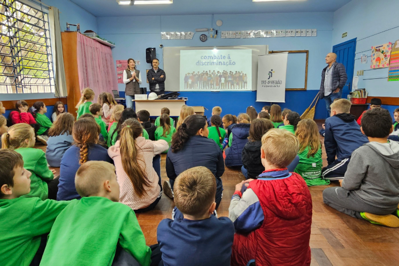 Sala de aula cheia de crianças, com palestrantes à frente.