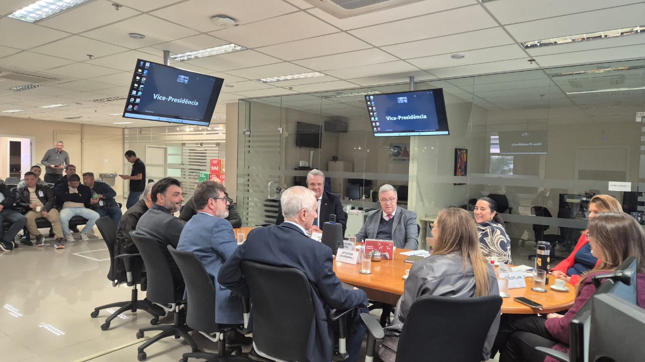 A foto mostra diversas pessoas sentadas ao redor de uma mesa. No lado esquerdo, algumas em pé.