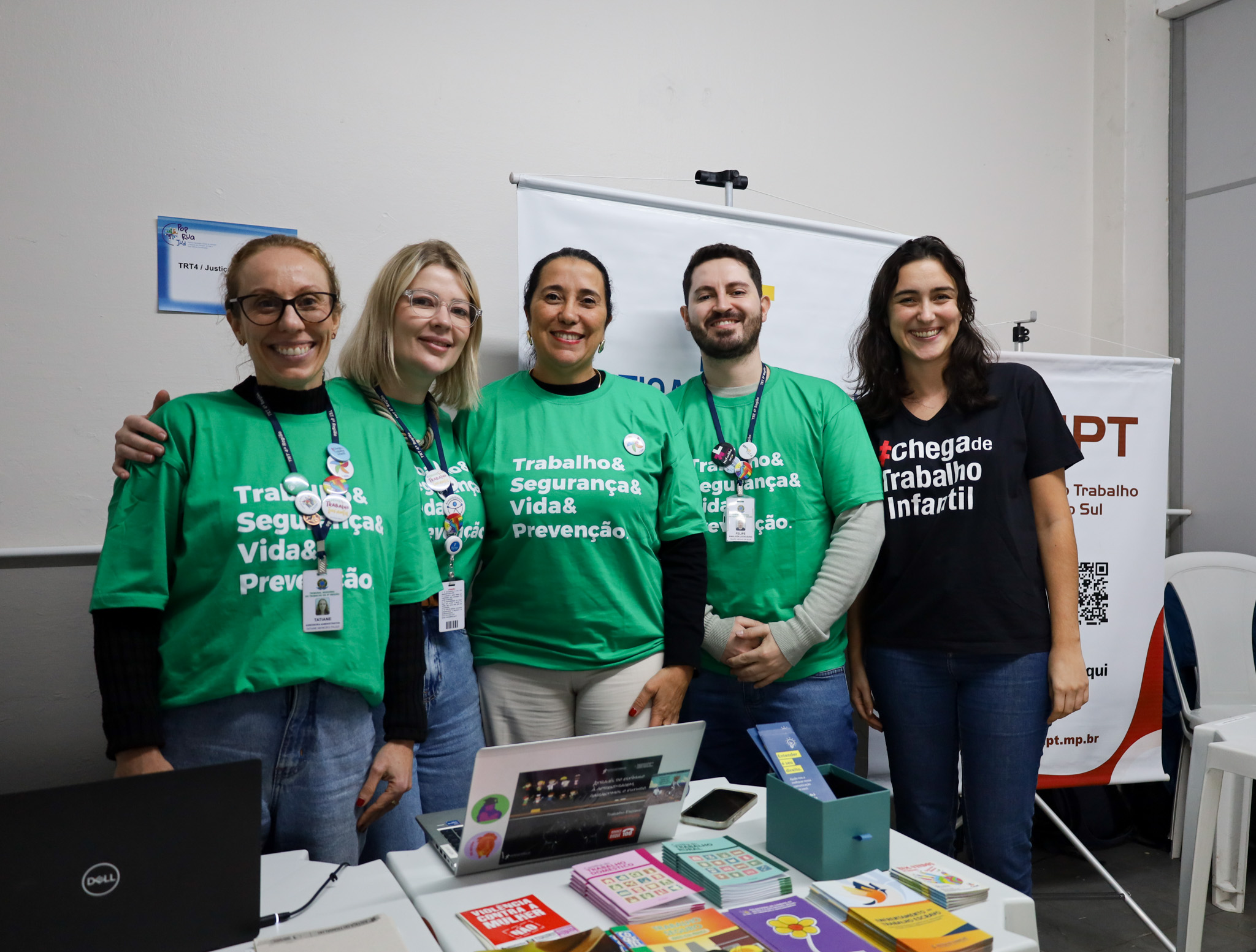 Cinco pessoas sorriem enfileiradas atrás de uma mesa. Quatro delas (três mulheres e um homem) vestem camisetas verdes com o texto "Trabalho & Segurança & Vida & Prevenção" impresso em branco. A mulher na direita veste uma camiseta preta com a frase "#chega de Trabalho Infantil". Sobre a mesa branca, há um notebook, um celular e vários panfletos e cartilhas coloridas. Ao fundo, destacam-se paredes claras e banners informativos da ação.