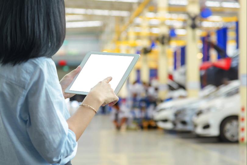 Em ambiente onde vários carros estão dispostos lado a lado, mulher verifica tablets. Ela está de costas, tem cabelos lisos e pretos e veste camisa azul claro.