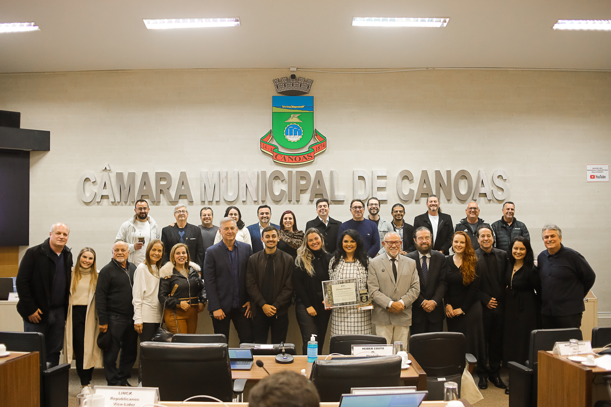 A foto mostra a desembargadora Tânia segurando um quadro da homenagem e vários convidados. Todos posando pra foto.