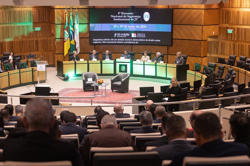 A fotografia mostra o plenário do TRT-RS, visto de cima, e ao longe, a mesa de abertura do evento com os magistrados e a magistrada sentados. Ao fundo, há um telão mostrando o banner do evento.