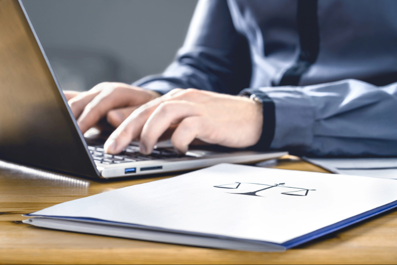 Homem digita em notebook. Ele usa camisa azul escuro. Sobre a mesa há uma pasta com desenho de uma balança, símbolo da Justiça.