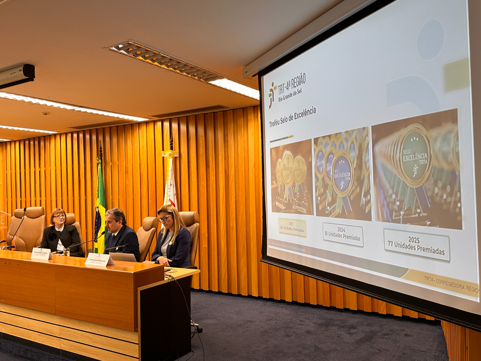 A foto mostra Telesca, Adolfo e Carolina apresentando projeto atrás de uma bancada com um telão ao lado.