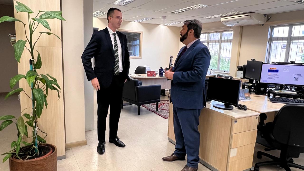 A foto mostra duas pessoas conversando lado a lado num ambiente fechado.
