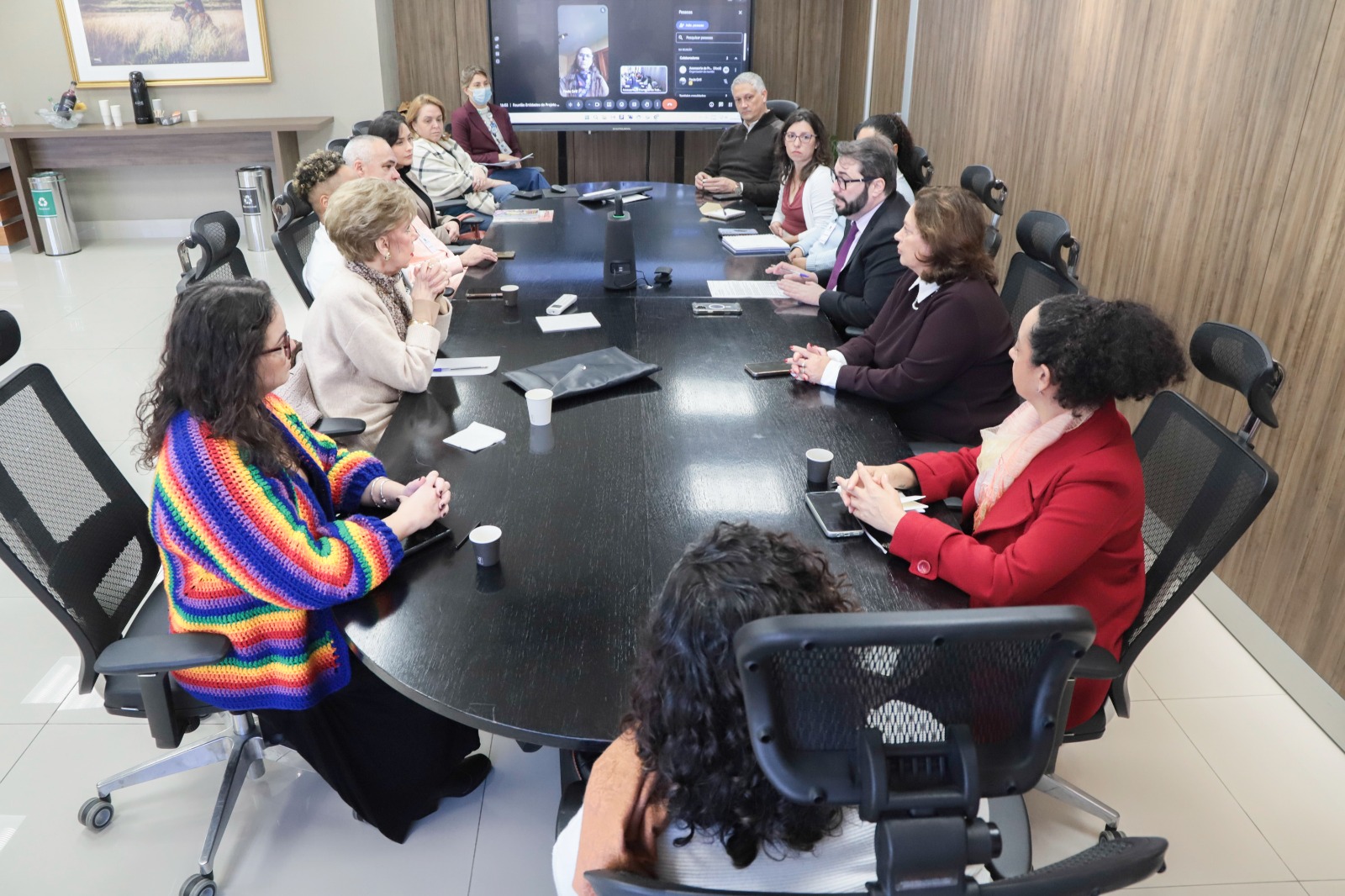 diversas pessoas sentadas em volta de uma mesa de reuniões. ao fundo há um telão ligado com alguém participando por videoconferência