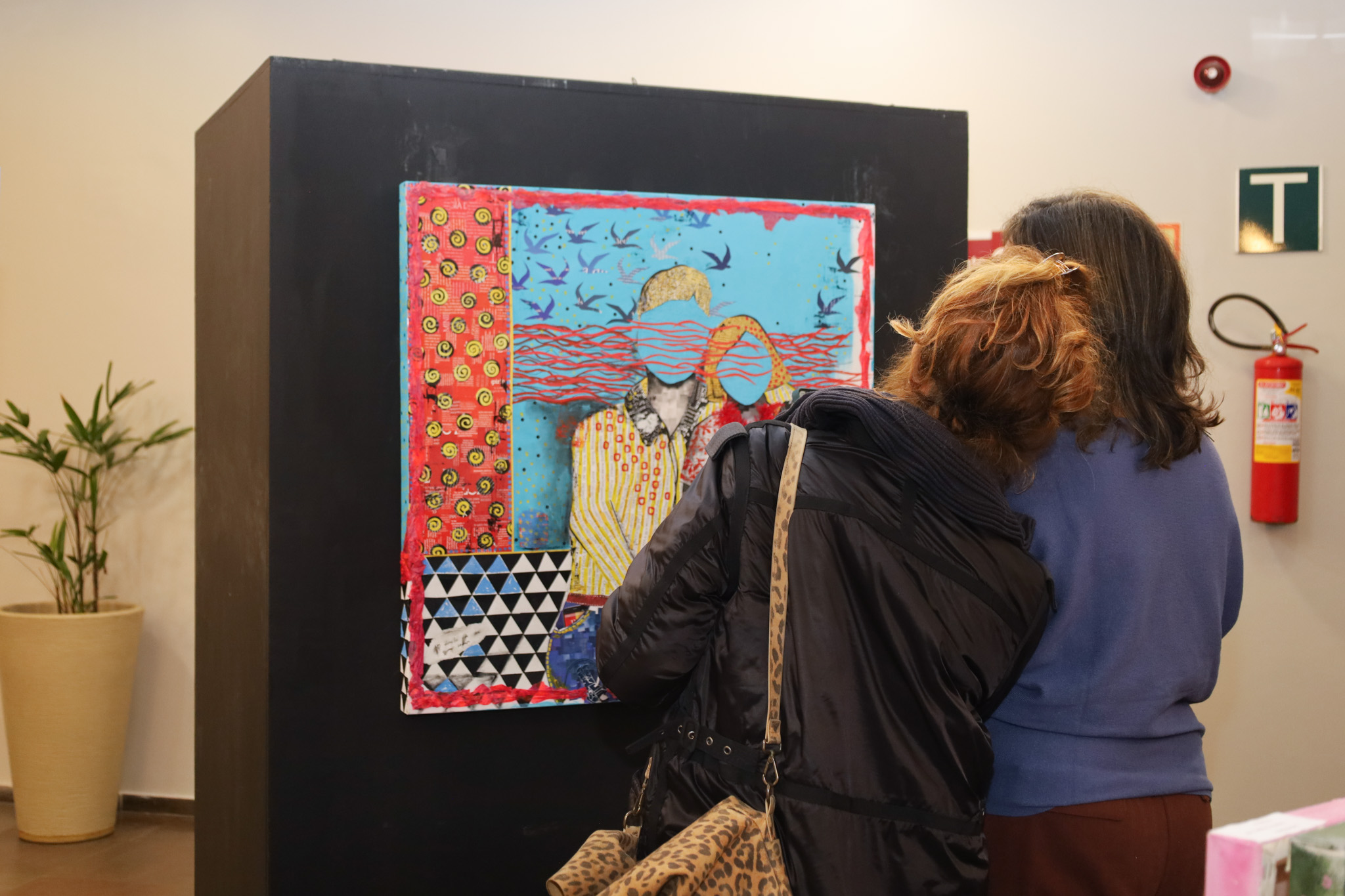 Duas mulheres, vistas de costas, admiram de perto uma obra de arte colorida fixada em um grande painel preto.