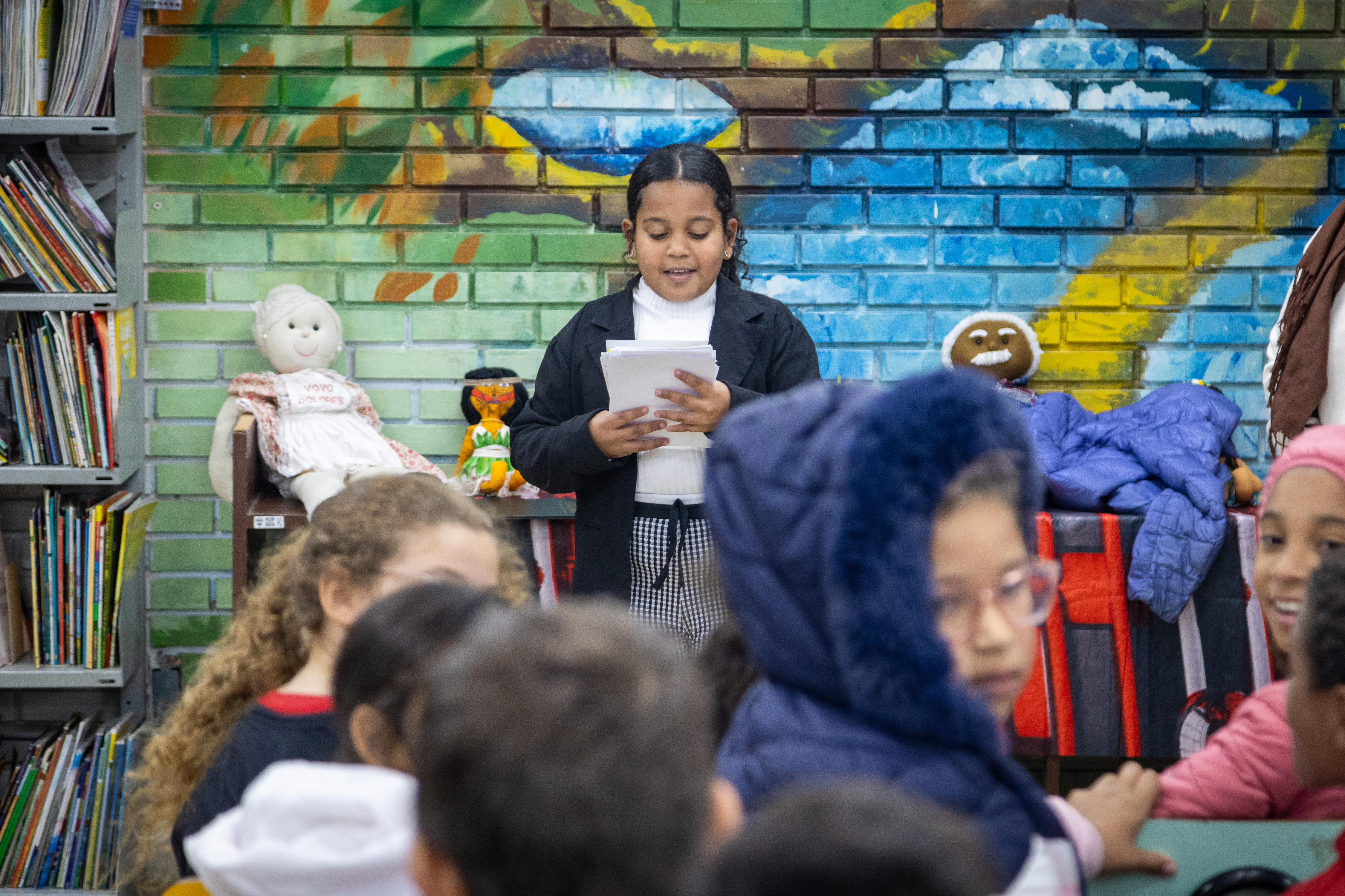 Aluna valentina lendo poema e crianças de costas na frente.