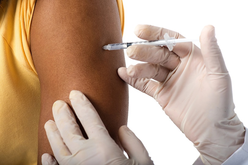 A foto ilustrativa mostra um profissional de saúde usando luvas descartáveis enquanto aplica uma injeção no braço de uma pessoa.