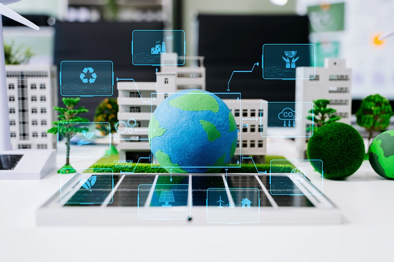 A imagem mostra uma representação conceitual de sustentabilidade e tecnologias ambientais. No centro, há um globo terrestre em miniatura sobre uma superfície que lembra painéis solares. Ao redor, aparecem maquetes de prédios, árvores e áreas verdes, formando uma pequena cidade sustentável.