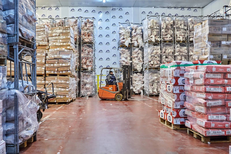 A fotografia mostra um frigorífico com caixas e pacotes de alimentos empilhados, e no centro da imagem há um homem pilotando uma empilhadeira.