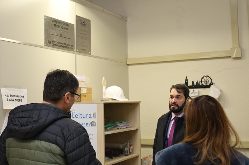 A fotografia mostra o presidente e a comitiva em sala do arquivo, e em destaque está a placa que mostra o nível da água na enchente, com aproximadamente 2 metros de altura.