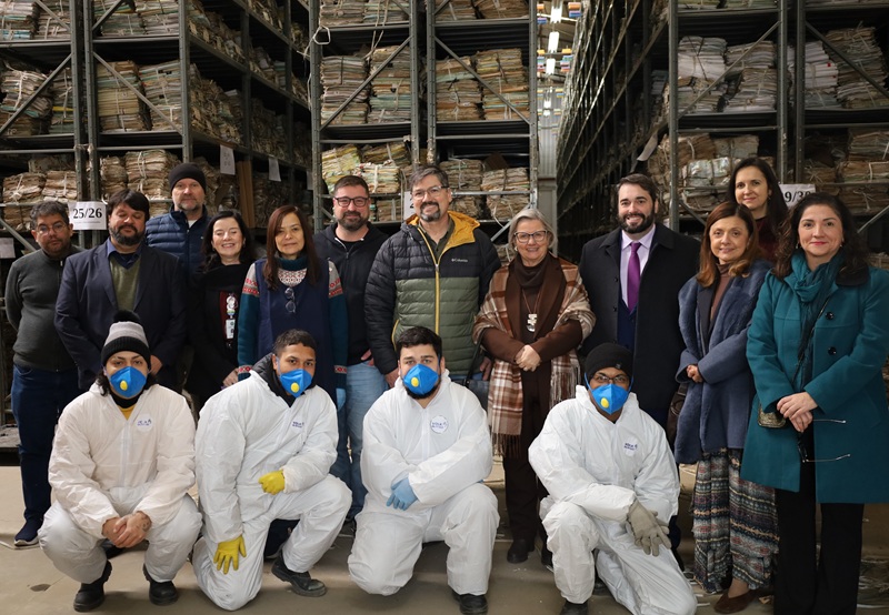 A fotografia mostra o presidente, sua comitiva, e parte da equipe que trabalha na recuperação dos processos no arquivo, posando de costas para as prateleiras de processos.