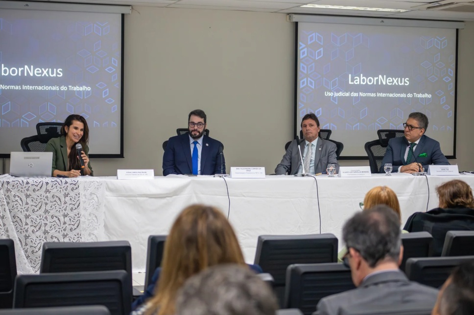 Foto da mesa de de autoridades durante apresentação da Labornexus. A juíza Carolina está falando ao microfone. O desembargador Alexandre também compõe a mesa, ao lado de outras autoridades.