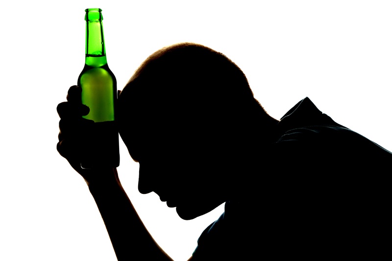 A fotografia mostra a silhueta de um homem, cabisbaixo, com uma garrafa de bebida alcoólica nas mãos.