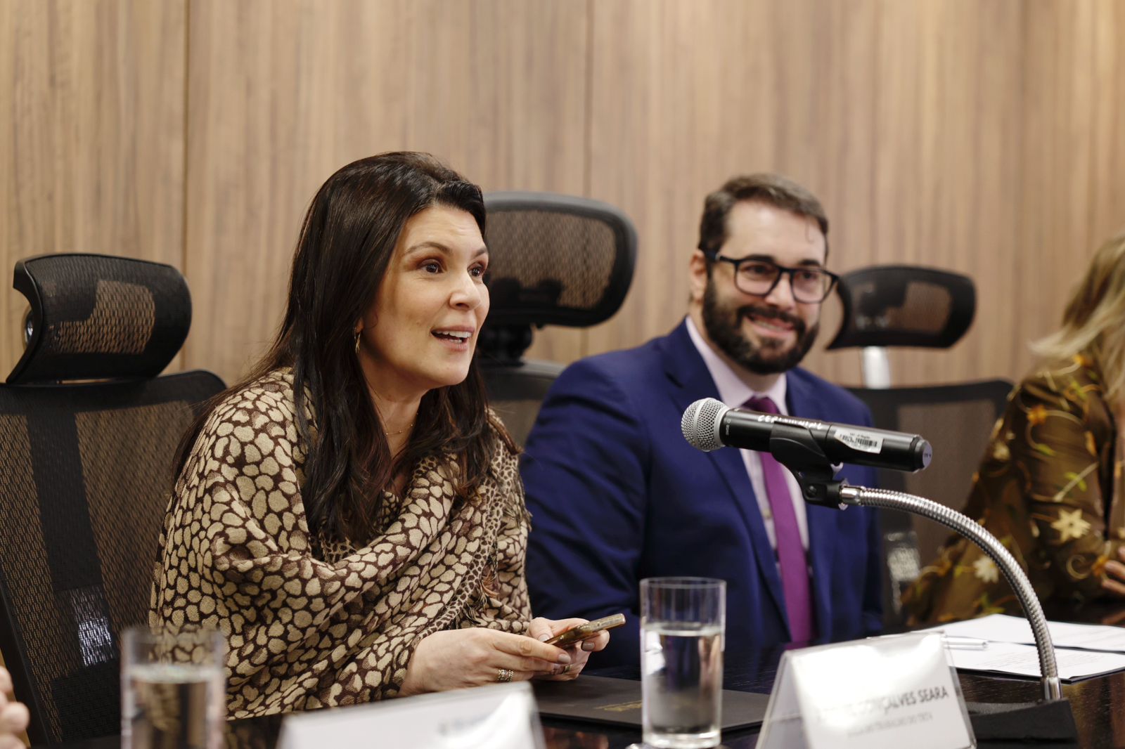 Raquel fala em primeiro plano, à esquerda. Ela é branca, de cabelo liso preto e comprido. Usa blusa marrom. O presidente está à direita da foto, sorrindo e olhando para o público. Ele é branco, de cabelo curto e escuro e barba escura. Usa terno azul, com camisa e gravata rosas.