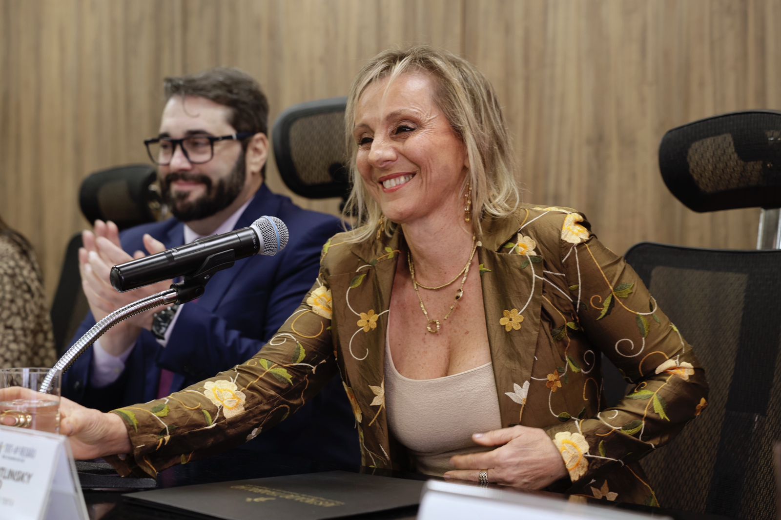 Ana Paula aparece em primeiro plano, na direita da imagem, sorrindo. Ela é branca e loira. Usa blazer marrom com estampas floridas. Ao fundo, o presidente aparece aplaudindo.