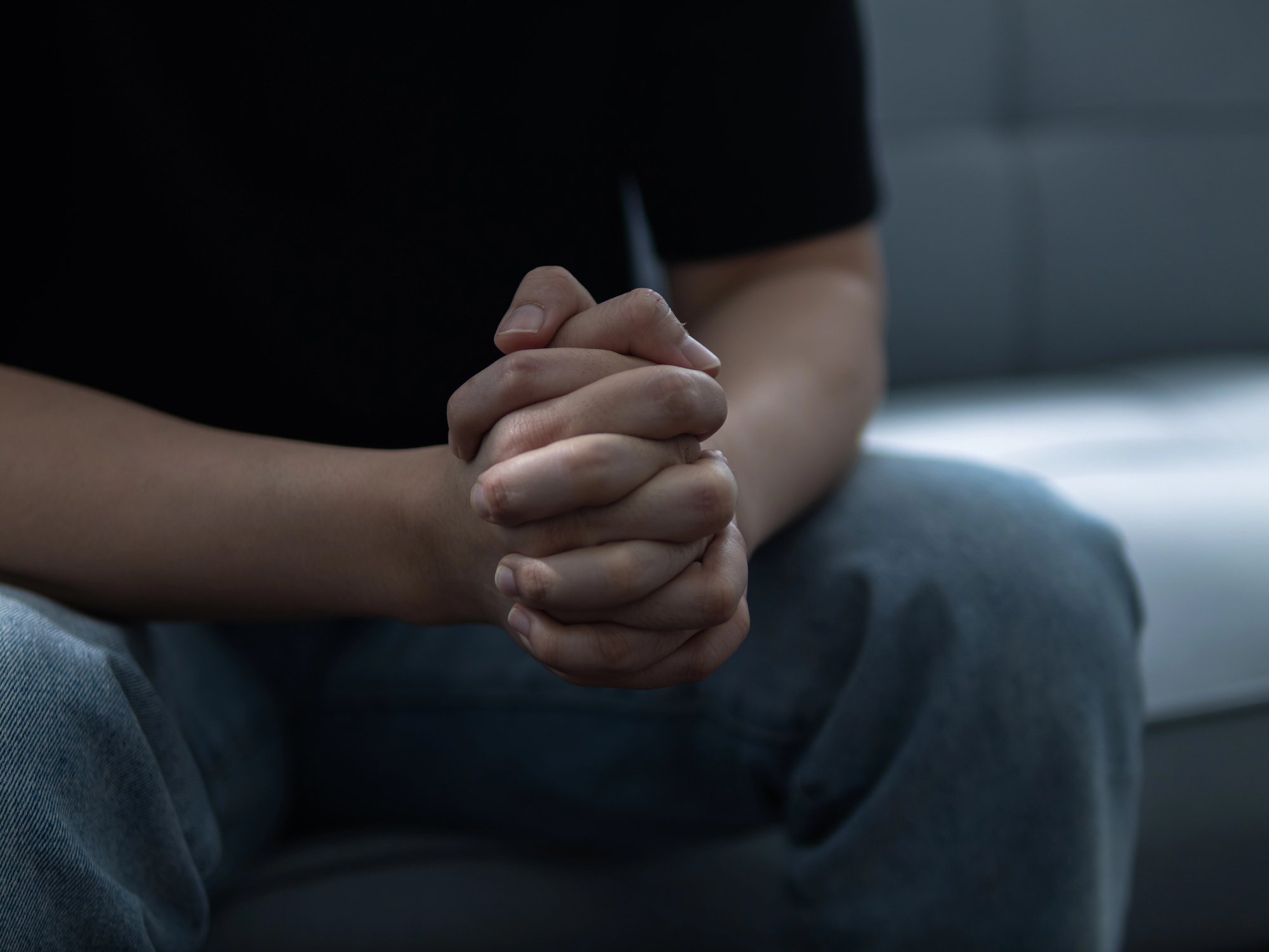 Pessoa sentada com as mãos fortemente entrelaçadas sobre o colo. O ambiente tem tons escuros e melancólicos, sugerindo um momento de reflexão, ansiedade ou profunda preocupação.