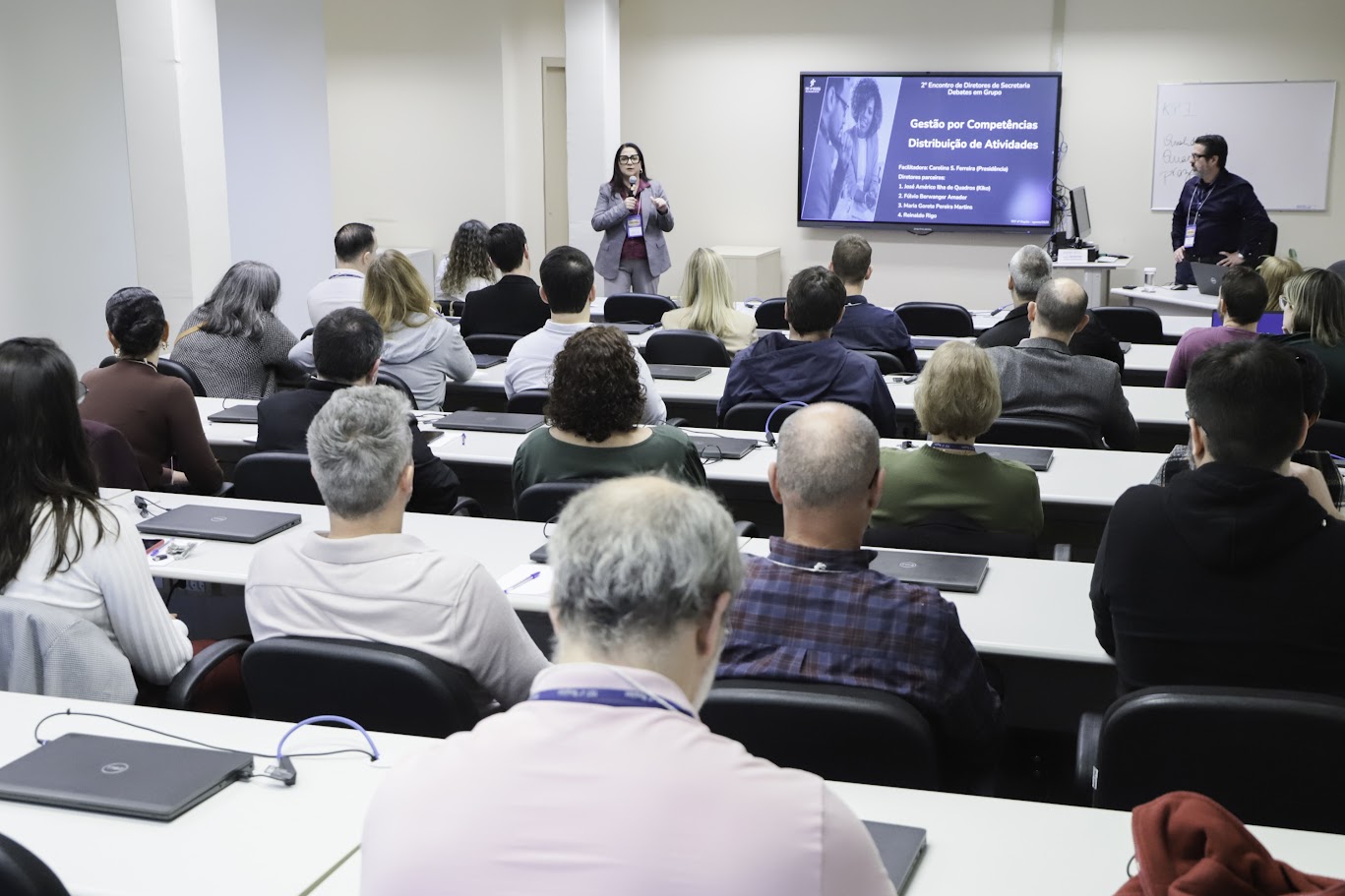 A fotografia mostra uma palestra, realizada no 2º Encontro, em 2025. Os diretores estão sentados em mesas de estudo, de costas para o fotógrafo. Na frente da plateia, dois palestrantes estão de pé