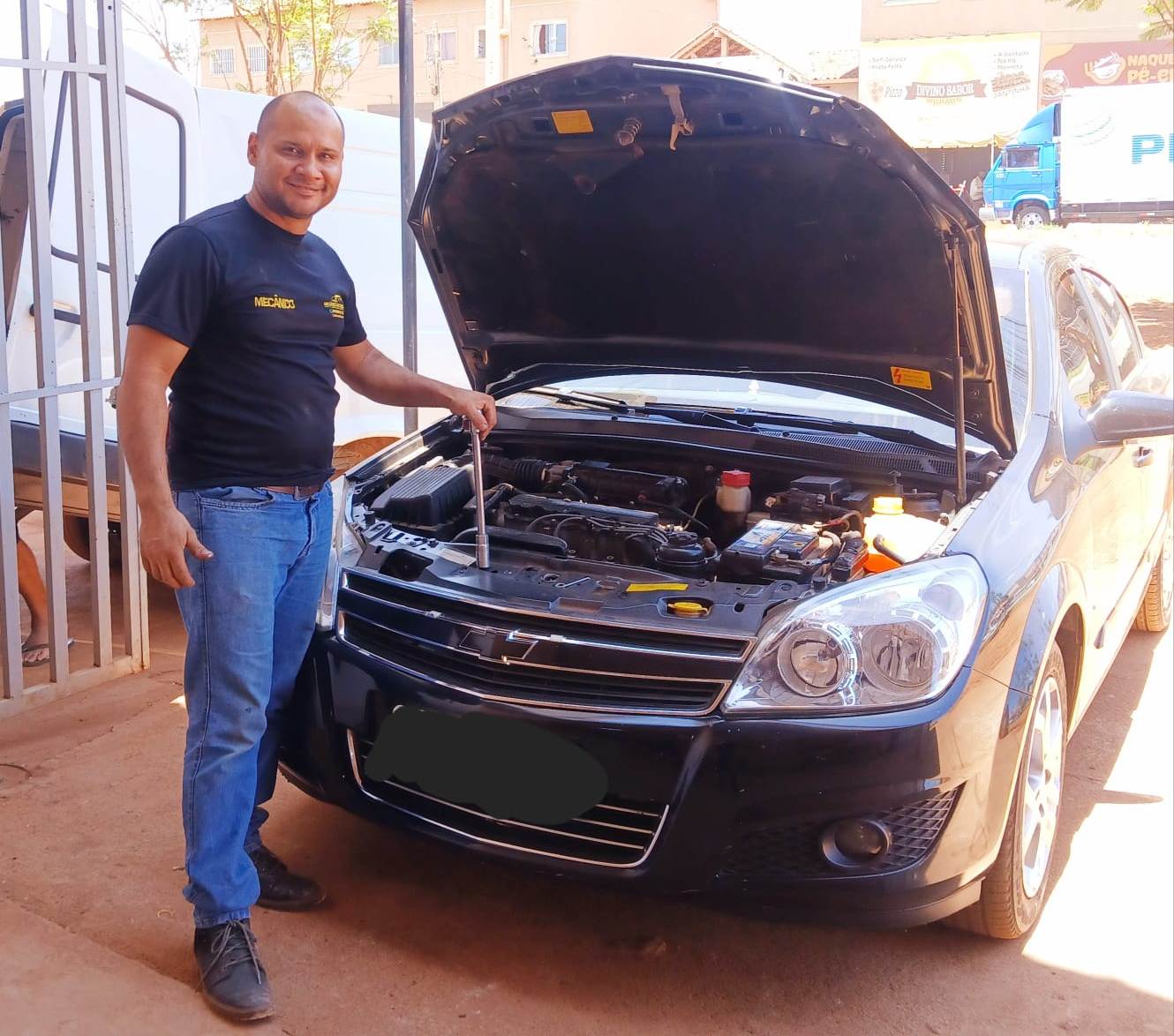 A imagem emilio_venezuelano_no_brasil.jpg mostra um homem sorridente ao lado de um carro preto com o capô aberto. Ele veste camiseta preta com a palavra "MECÂNICO" estampada em amarelo, calça jeans azul e sapatos escuros. Com a mão esquerda, ele segura uma ferramenta longa apoiada no compartimento do motor, que está exposto. O cenário é ao ar livre, em solo de terra batida, próximo a um portão de metal branco. Ao fundo, veem-se construções urbanas e um caminhão azul.