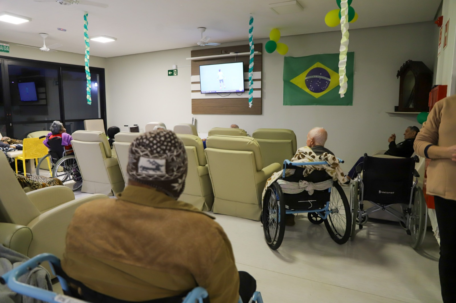A foto mostra idosos de costas vendo TV.