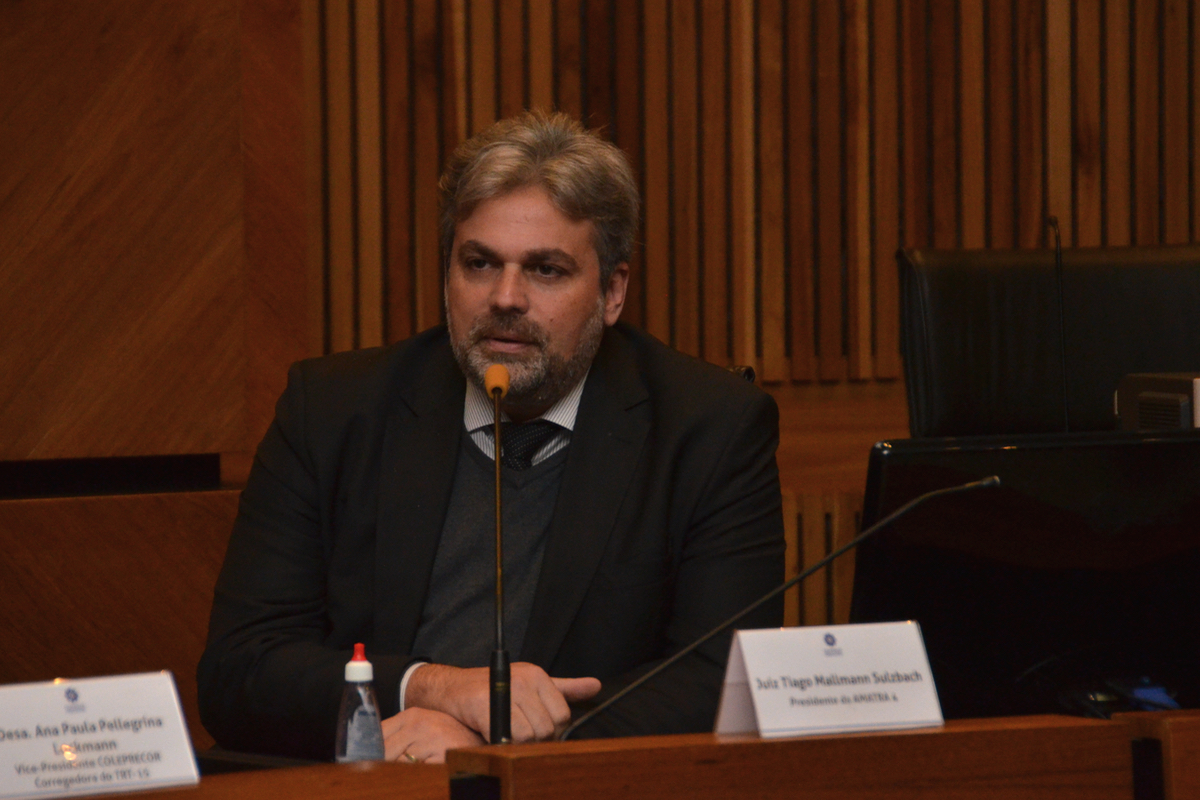 Tiago Sulzbach é branco, grisalho, usa terno escuro e blusão cinza. Fala ao microfone.