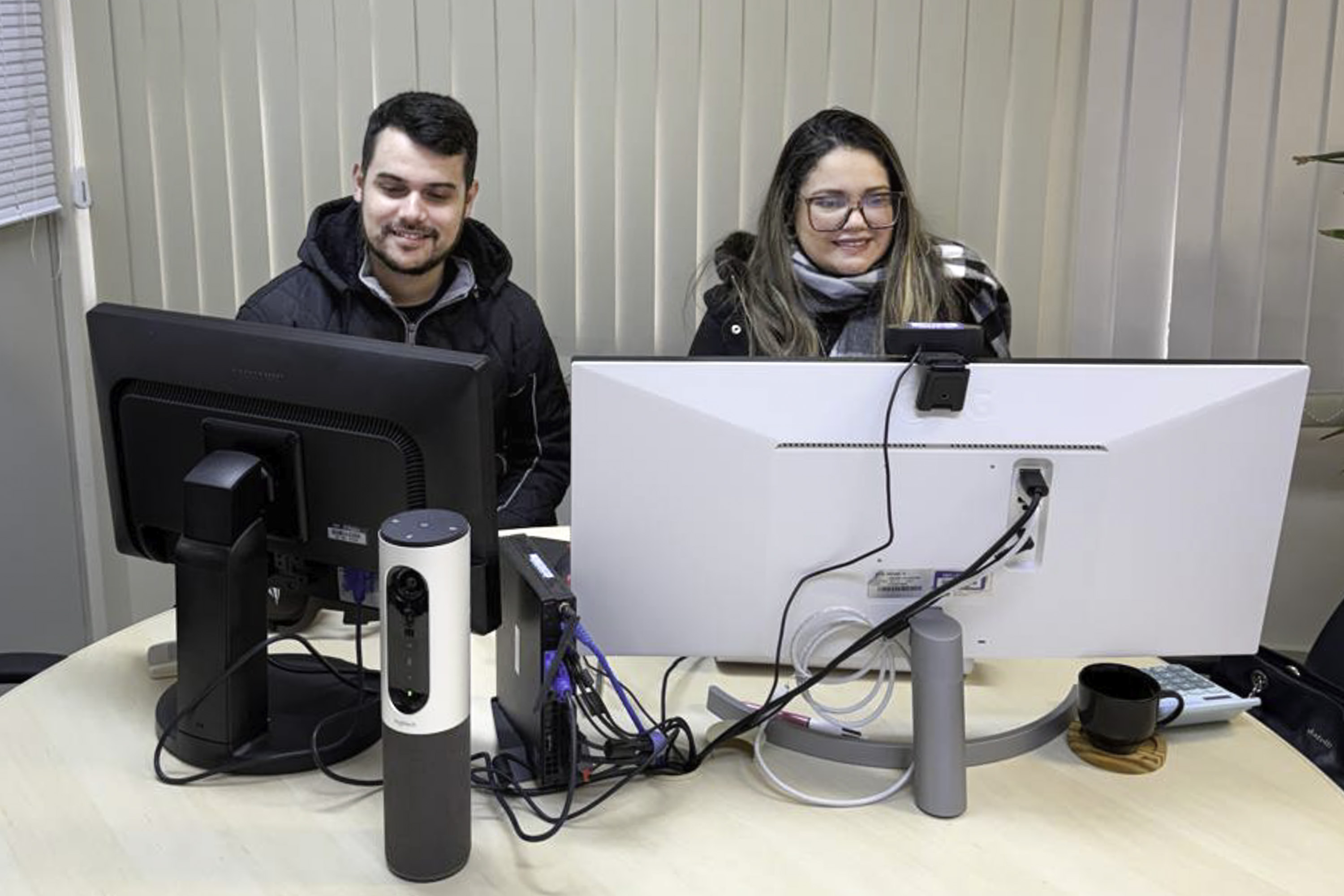 Gabriel e Joana sorriem. Estão em uma mesa redonda de conciliação, um ao lado do outro. Cada um olha para a tela de um computador. 