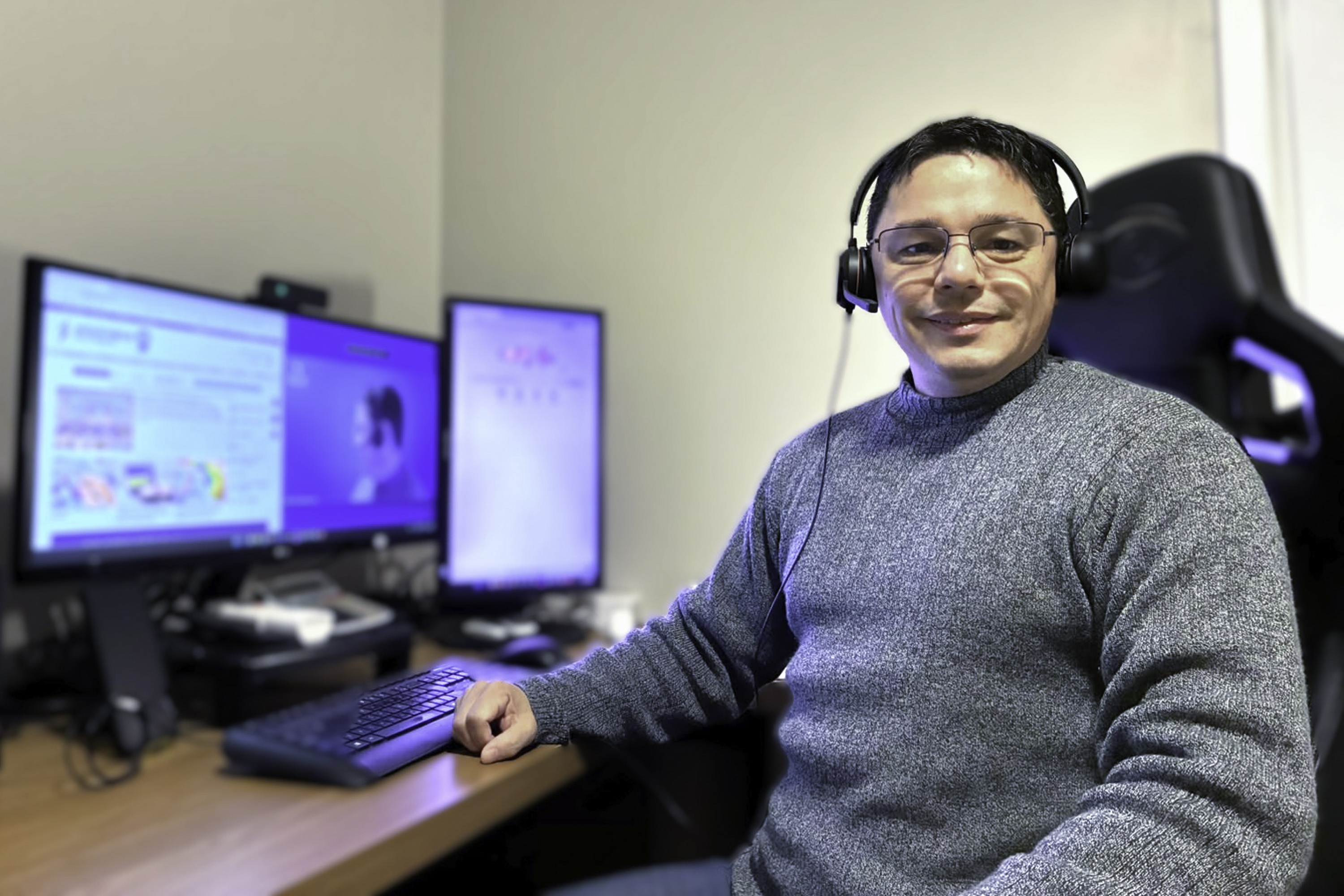 João usa um fone de ouvidos e está sentado em um cadeira, sorrindo e olhando para a câmera. a seu lado aparece a mesa de trabalho com um computador ligado. 