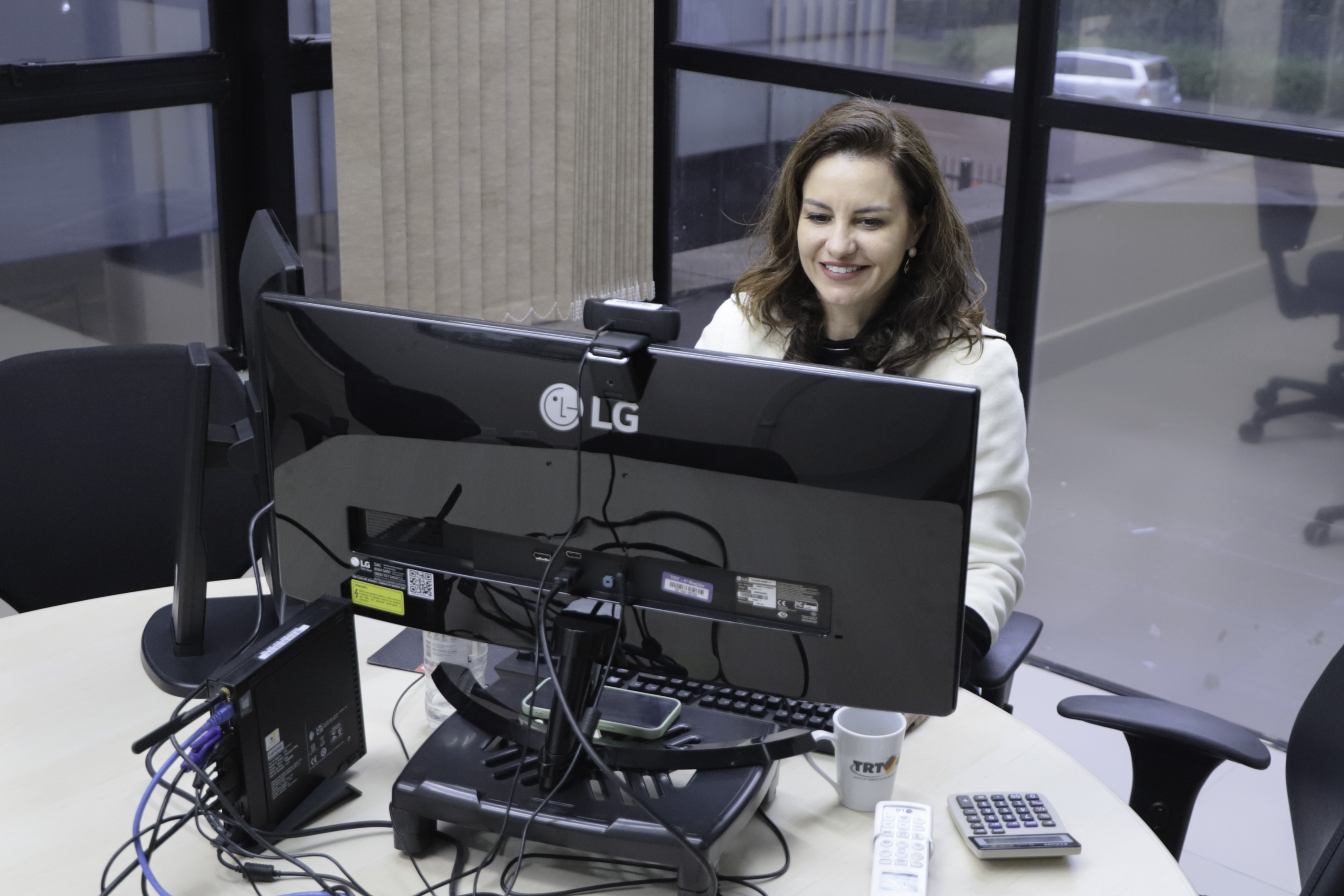 Gabriela sorrri olhando para a tela do computador. ela trabalha em uma mesa redonda e Atrás delas aparecem janelas abertas. 