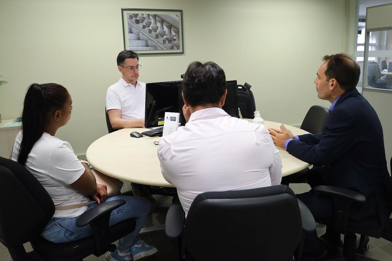 Duas pessoas aparecem de costas e uma de perfil em primeiro plano. A de perfil fala e gesticular. No fundo, ao centro, um conciliador olha para a tela do computador. 