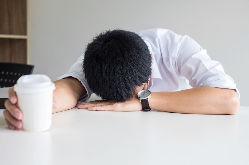 Foto ilustrativa de uma pessoa com a cabeça apoiada sobre uma mesa e segurando um copo de café descartável, aparentando cansaço.