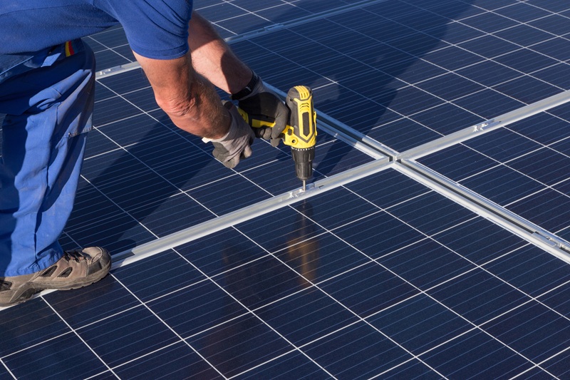 A fotografia mostra em close um trabalhador instalando placas solares com uma parafusadeira.