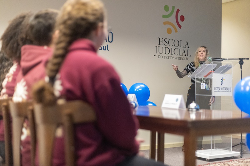 A fotografia mostra uma das equipes vista de costas e a servidora Renata falando no púlpito.