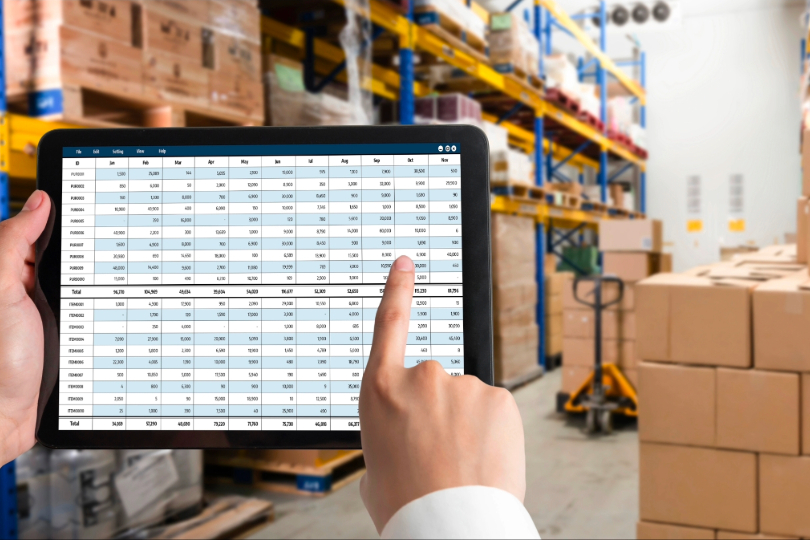 Mão masculina aponta planilhas em tela de tablet. Ao fundo, há prateleiras com caixas de mercadorias.