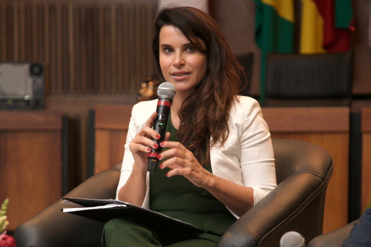 Carolina é branca, tem cabelo castanho liso e olhos claros. Está sentada em uma poltrona e fala ao microfone. Usa blusa preta e blazer branco. Ao fundo aparecem bandeiras da Justiça do Trabalho, do Brasil e do Rio Grande do Sul.