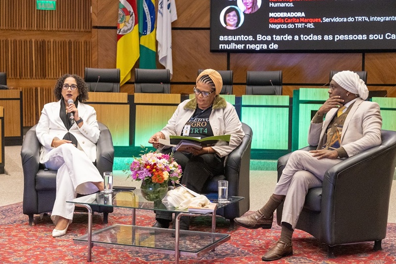 Na imagem, Karla está sentada falando ao microfone, ao lado de Gladis e Gleidson, também sentados.