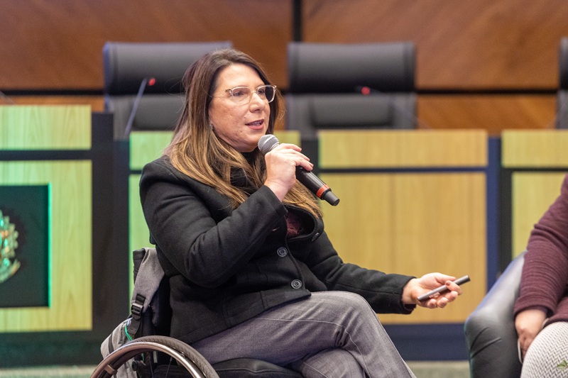 Na imagem, Ekaterini está falando ao microfone e usa cadeira de rodas.