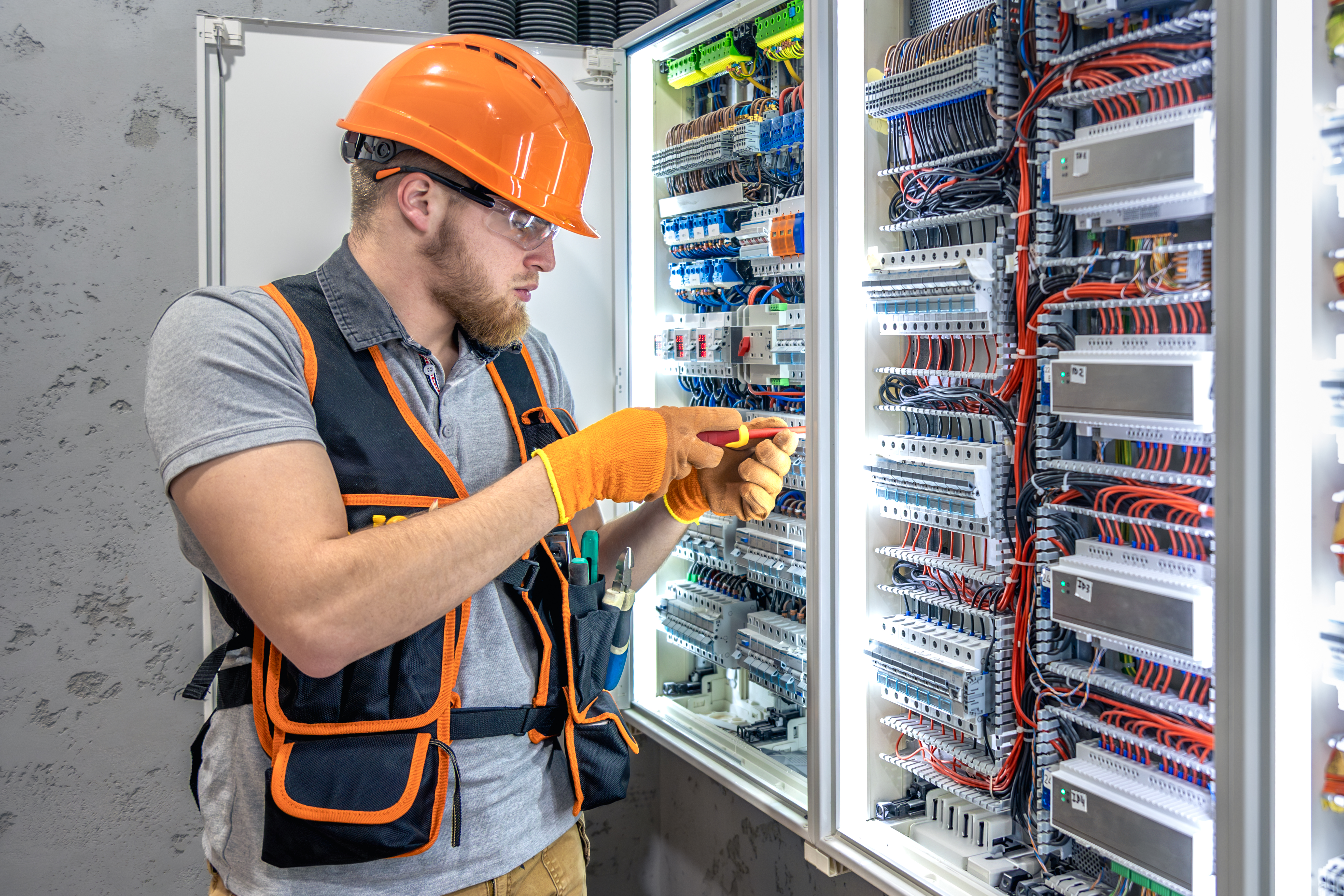 Imagem de um homem parafusando um quadro de luz. Usa óculos de proteção, luvas e colete.